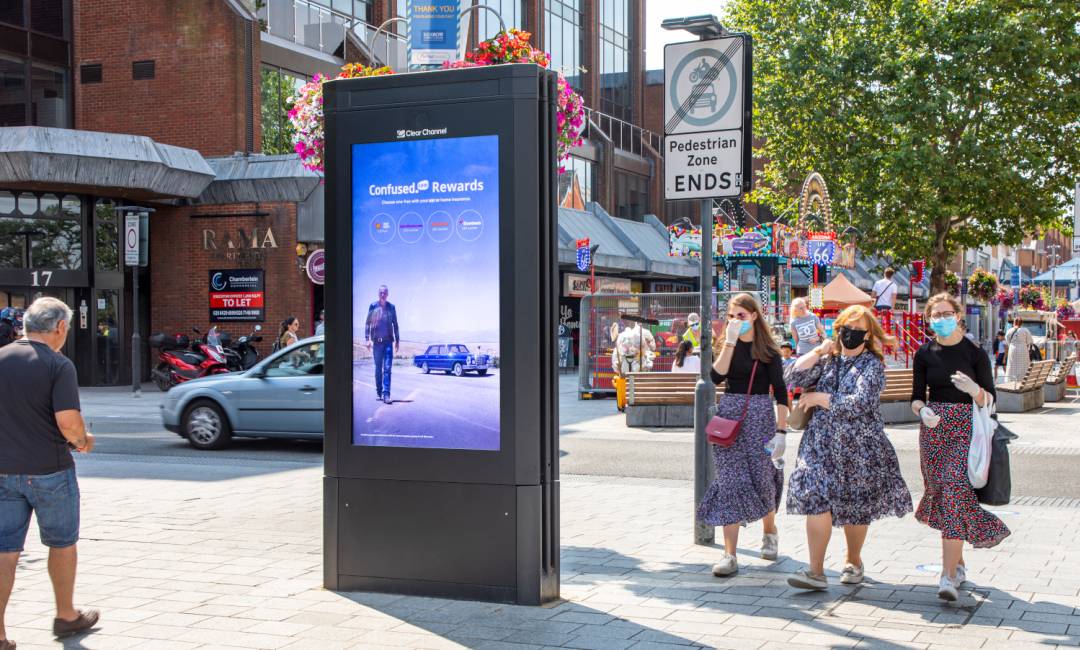 People walking past an Adshel Live display in the middle of a busy square during the day