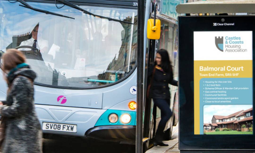 People in front of a digital bus stop screen featuring a campaign by Castles & Coasts Housing Association