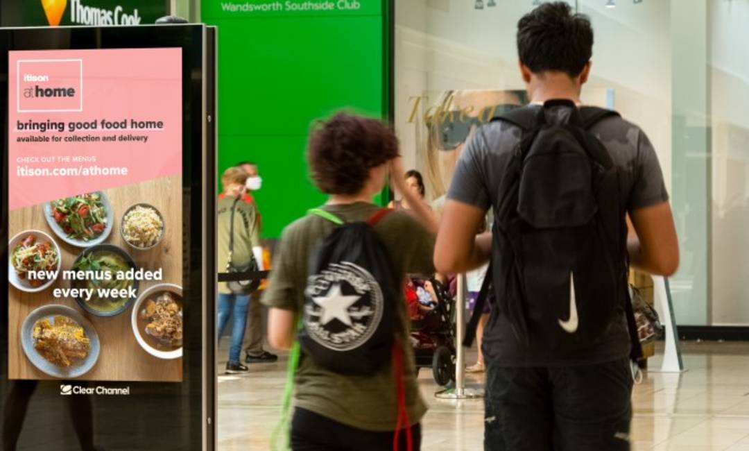 A shopping mall digital screen featuring a food campaign with people in Covid-19 masks walking by