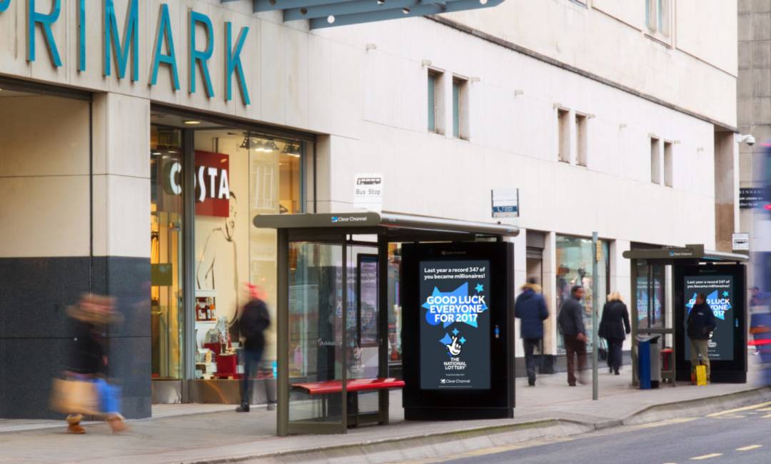 National Lottery advertisement at a bus shelter by a Primark as people walk by during the day