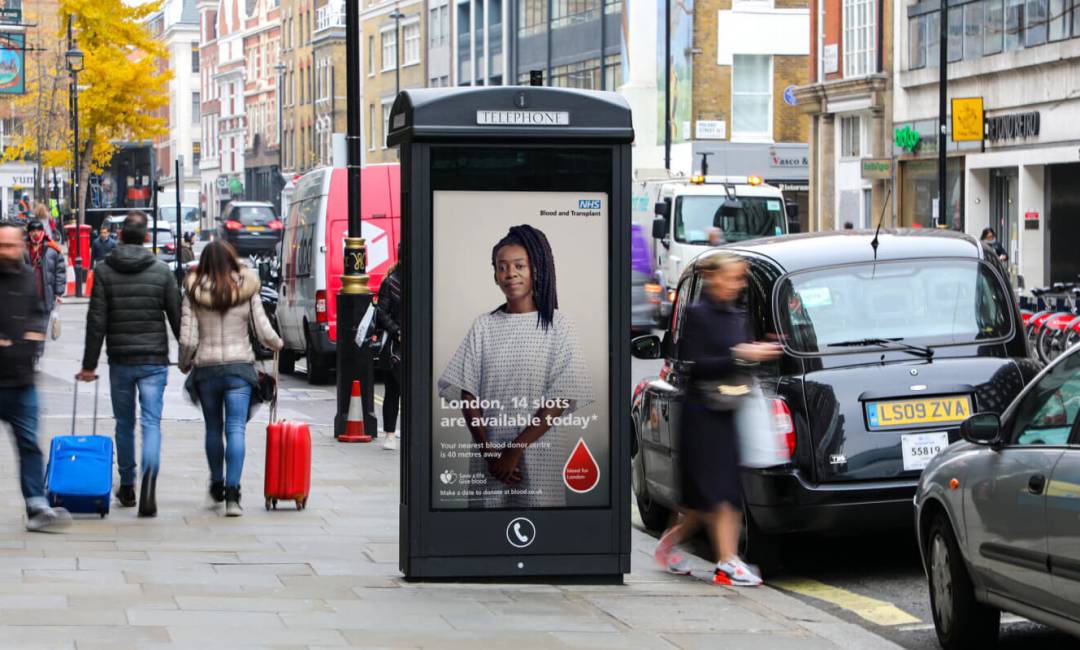 A digital advertising screen on a phone kiosk in London showing a dynamic ad for NHS Blood and Transplant