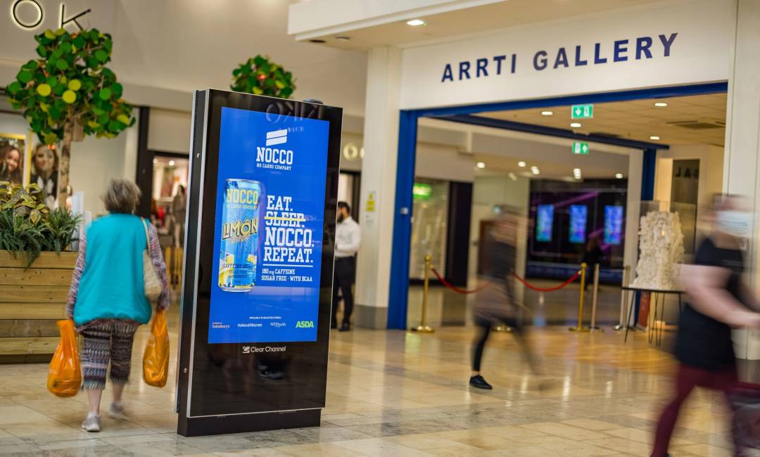 A Nocco drink ad on a digital advertising screen inside a shopping mall with people walking past