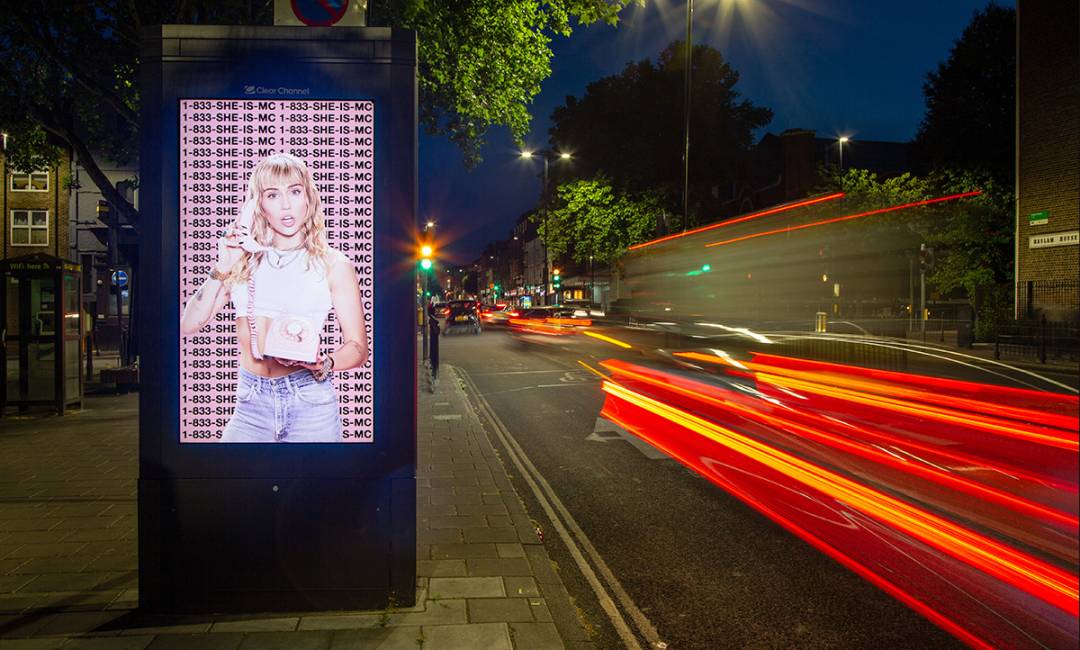 An Adshel Live screen on a free standing roadside unit at night showing an ad for a Miley Cyrus album