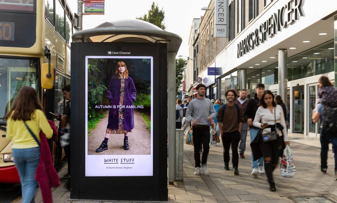 Pedestrians walking down the street during the day past a bus shelter with a White Stuff advertisement on its side