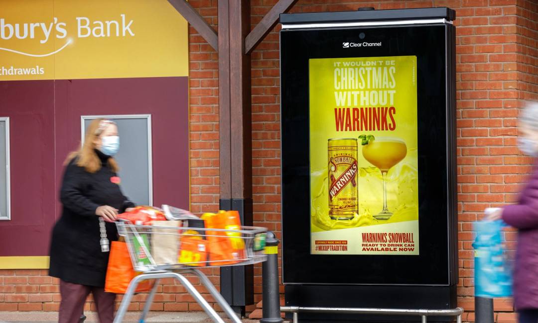 A Warninks ad on a digital screen outside a Sainsbury's supermarket with a shopper walking past
