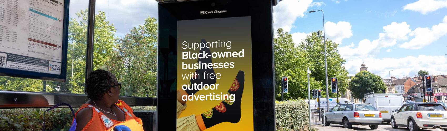 Black woman sat at a bus stop with digital advertising poster saying 'supporting black-owned businesses with free outdoor advertising'