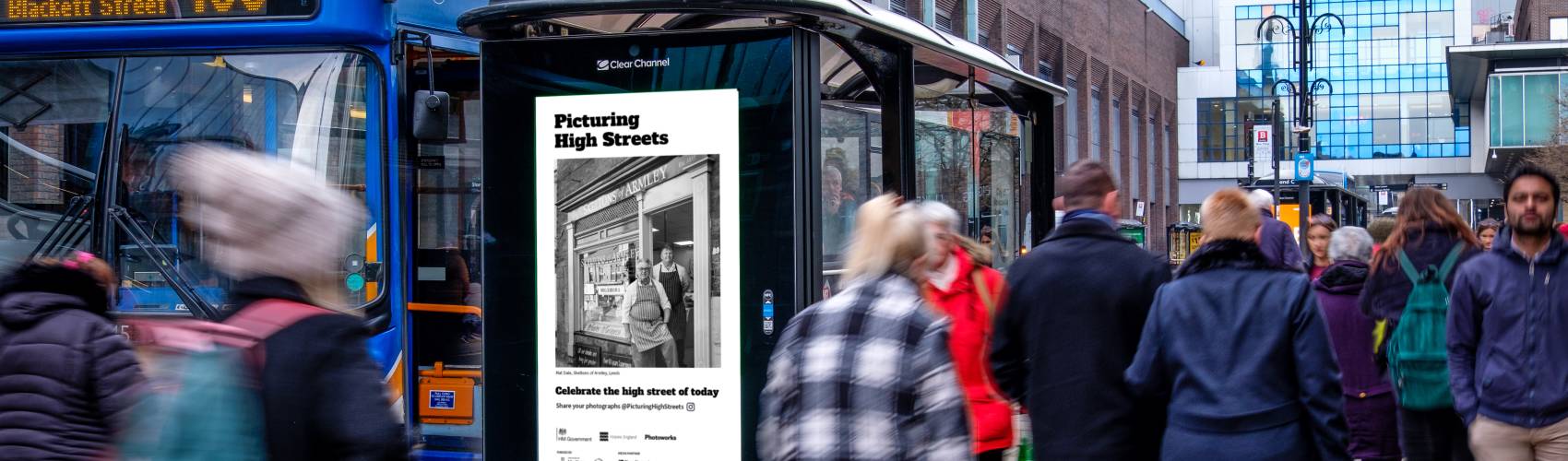 Digital bus shelter screen in Newcastle next to a static bus and a lot of people walking