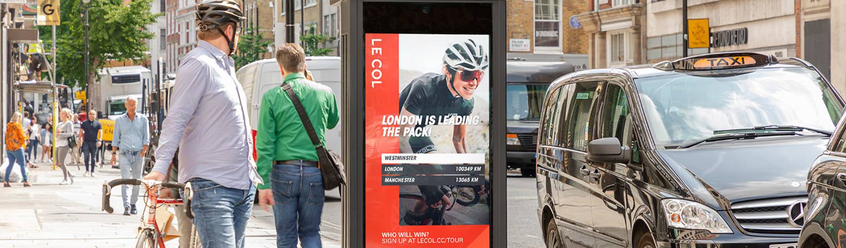 A cyclist on the pavement looking at a Le Col cycle campaign on an Adshel Live panel