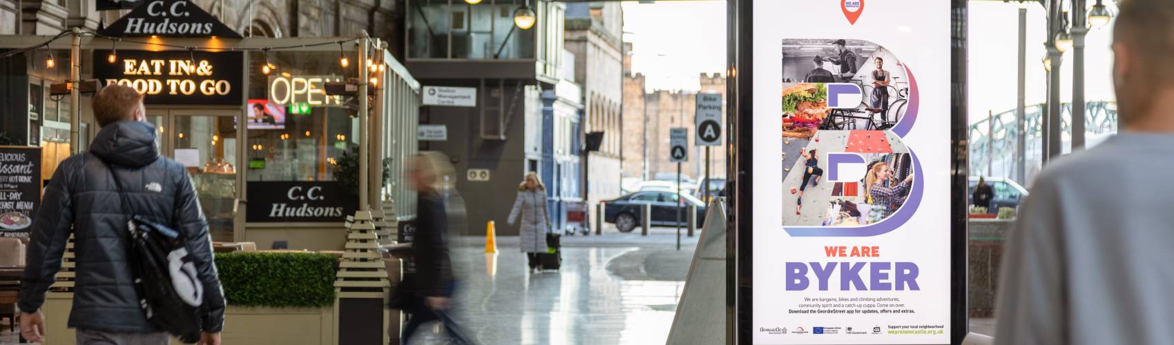 A Adshel live screen inside a train station with people walking past.