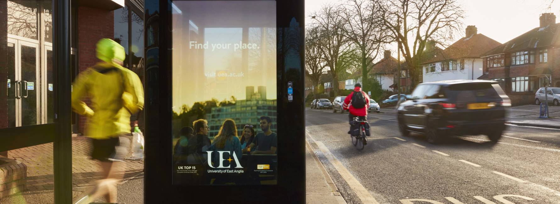 UEA's Bus Shelter ad as people run and cycle past