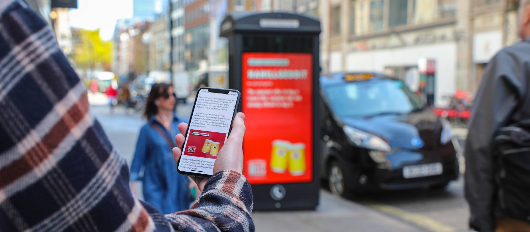 Person holding mobile phone with a beer advert on screen and a digital outdoor advertising panel with the same brand in the distance