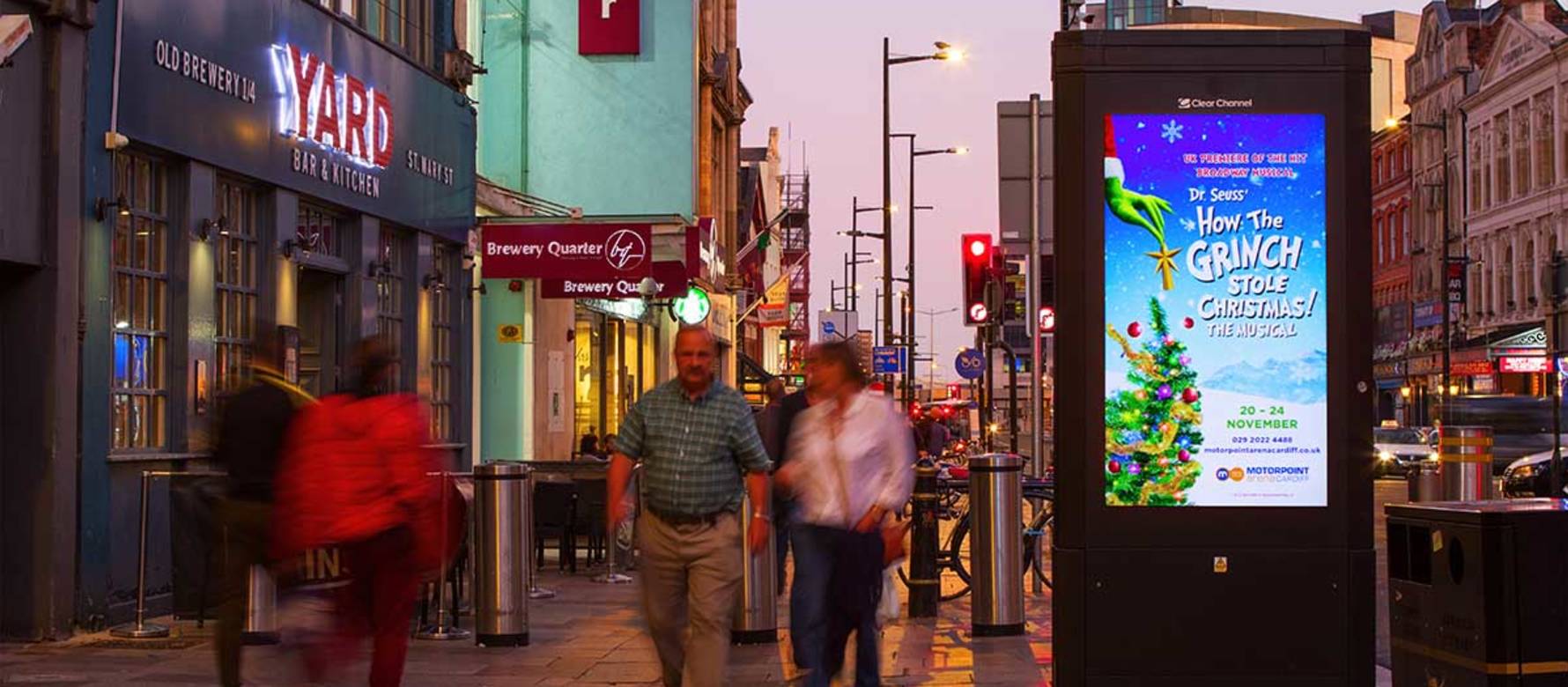 People walking past an Adshel Live display, featuring an advertisement for How the Grinch Stole Christmas, at night