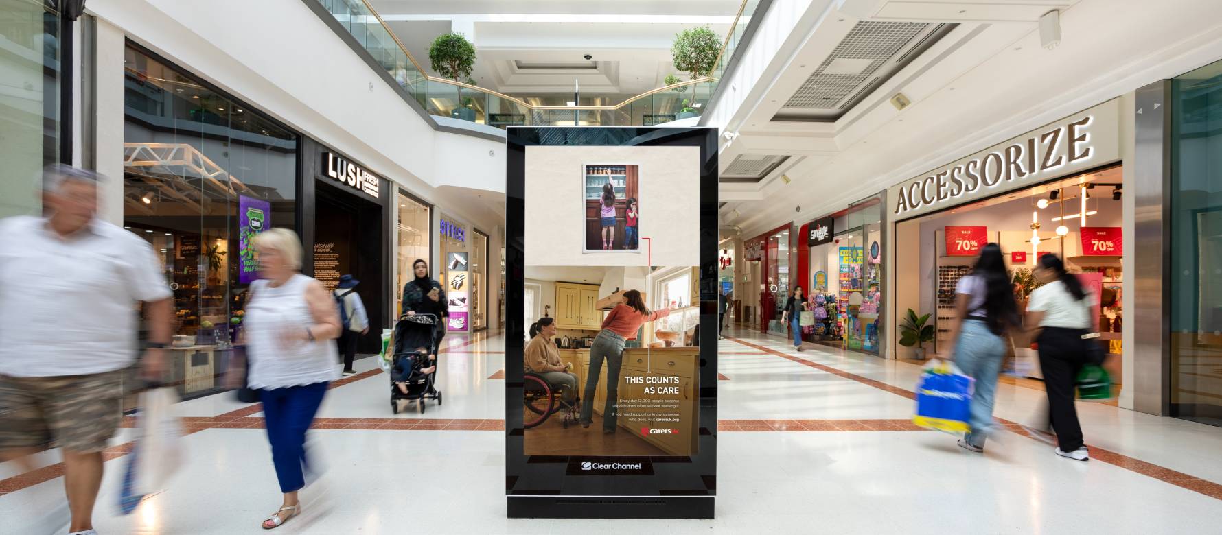 ‘This Counts As Care’ campaign advertisement for Carers UK featured on a Malls Live digital screen in a busy shopping center as individuals walk by in the middle of the day.