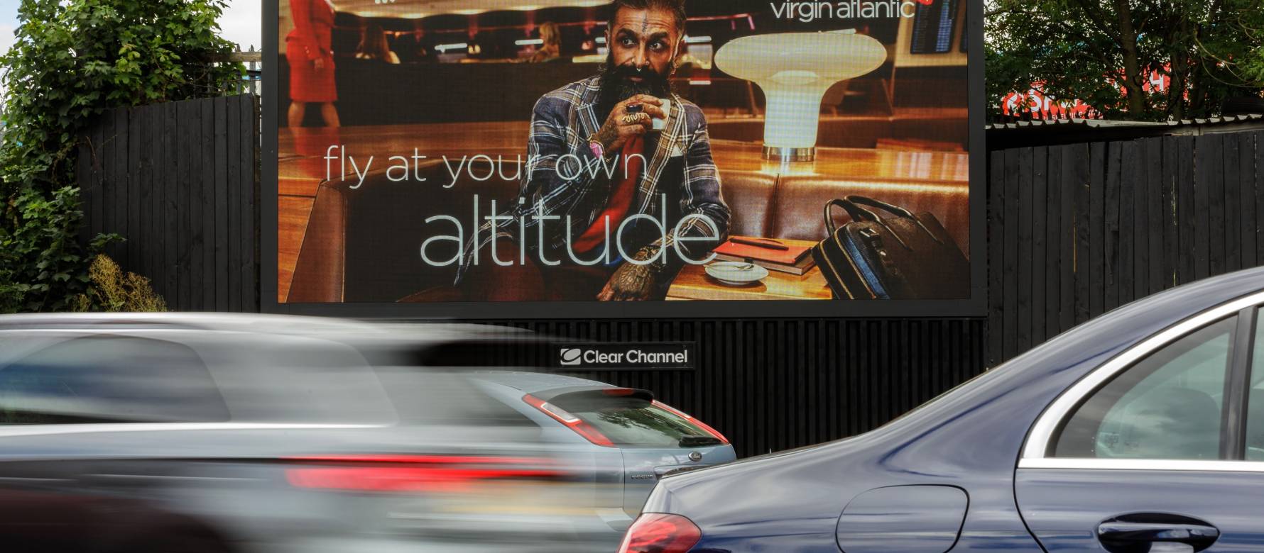 A Virgin Atlantic advertisement of a person drinking a coffee in a Virgin Atlantic airport lounge, on a Clear Channel billboard by a busy road