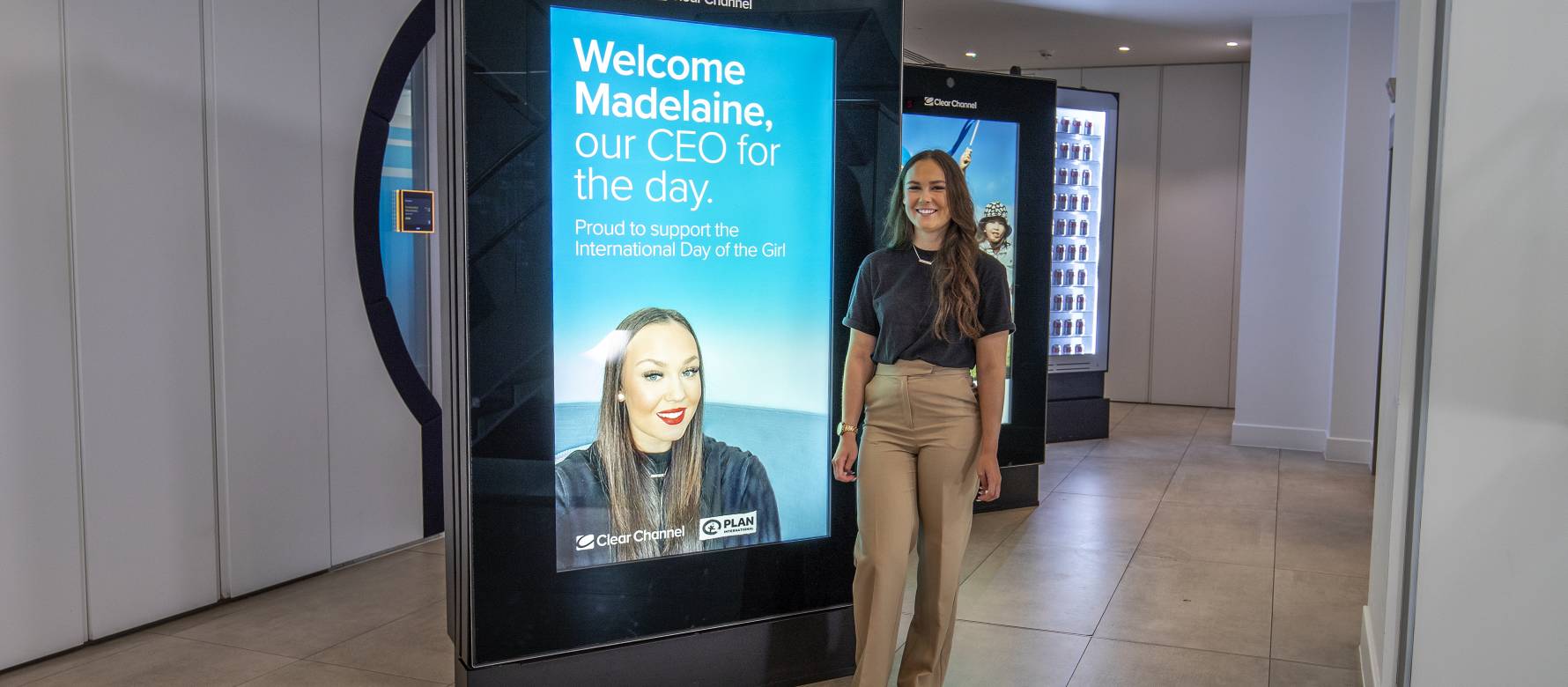 Madelaine in the reception of Clear Channels Golden Square office standing next to a digital display, that says welcome I'll see you home for the day, proud to support the International Day of the girl