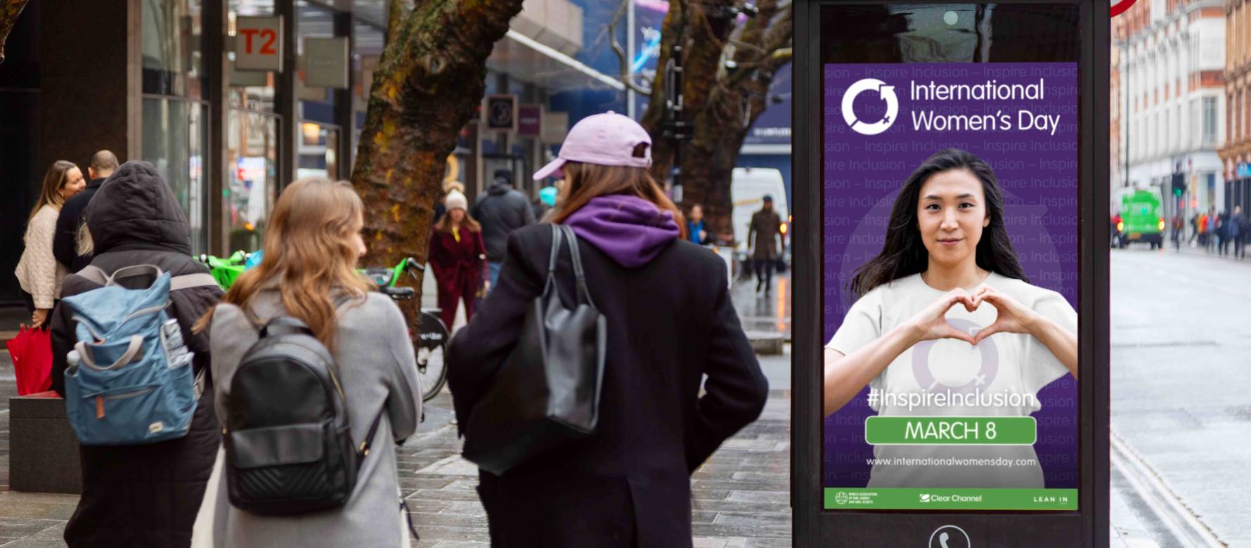 An international Women's Day advertisement displayed on the side of a New World Payphone on a busy street as pedestrians walk by