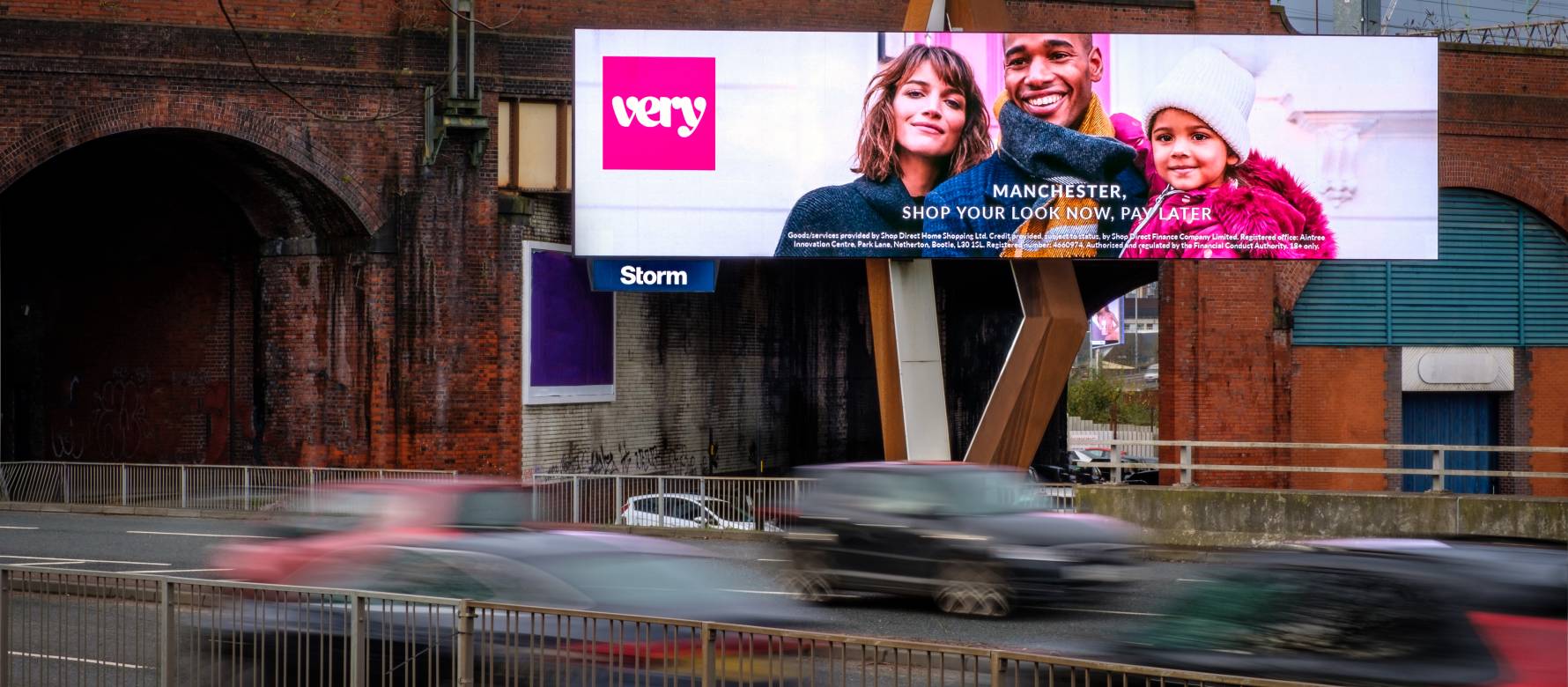 Billboard over busy road, beside train arch, displaying a fashion brand