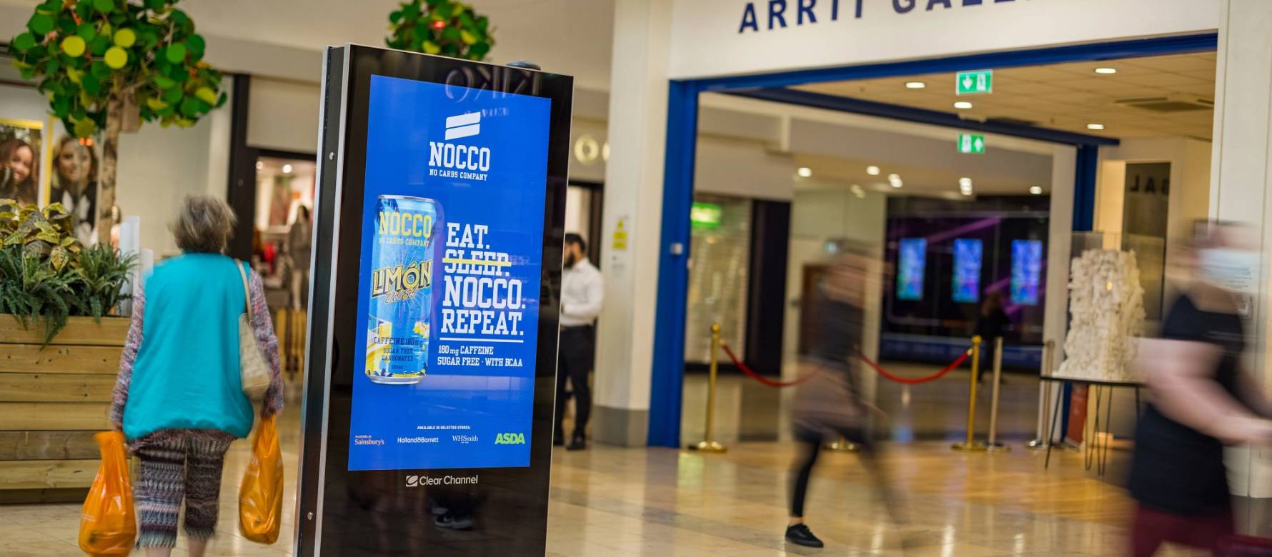 A Nocco drink ad on a digital advertising screen inside a shopping mall with people walking past