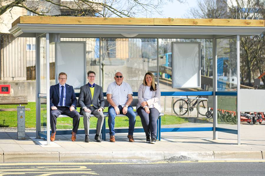 A group of people sitting on a bus shelter that has a living roof.