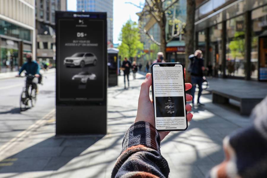 Hand with phone in showing a car ad that matches the one on the digital screen in the background