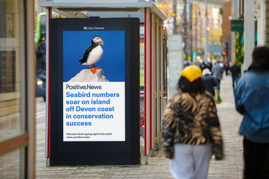 A Positive.News headline displayed on a Clear Channel UK Adshel Live display at a bus stop as people walk past during the day