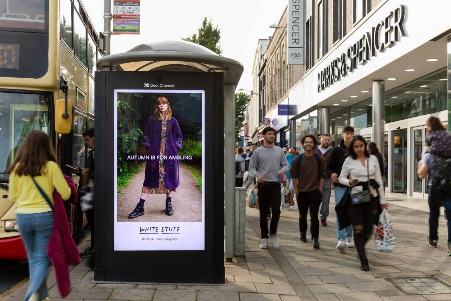 Pedestrians walking down the street during the day past a bus shelter with a White Stuff advertisement on its side