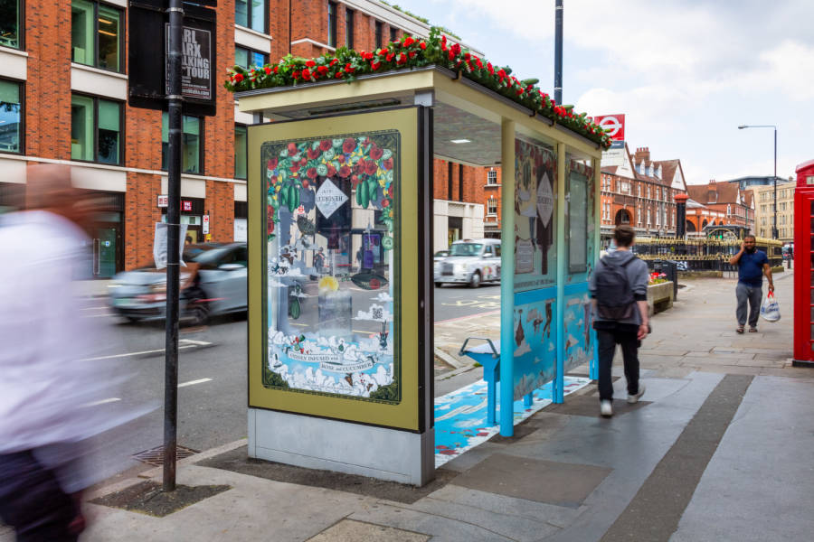 People walking past a bus shelter which features a 3D Hendricks advertisement on a busy street during the day