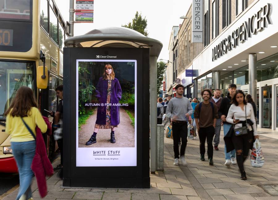 A poster advertising White Stuff clothing on an Adshel Live screen at a bus shelter on a busy high street.