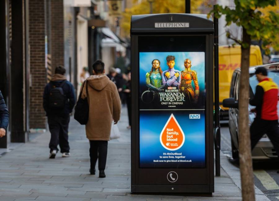 Digital pay phone panel with a Wakanda and NHS advert next to people walking on the pavement