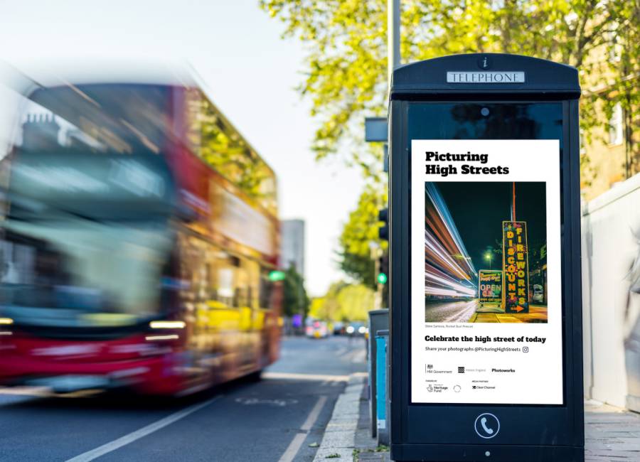 A New World Payphone featuring a Picturing High Streets competition winning image on its digital screen, during the day as a bus and pedestrians pass by