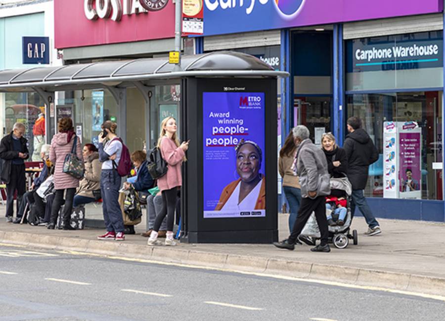 Lots of people waiting at a bus stop on a high street with a screen showing Metro Bank advertisement.