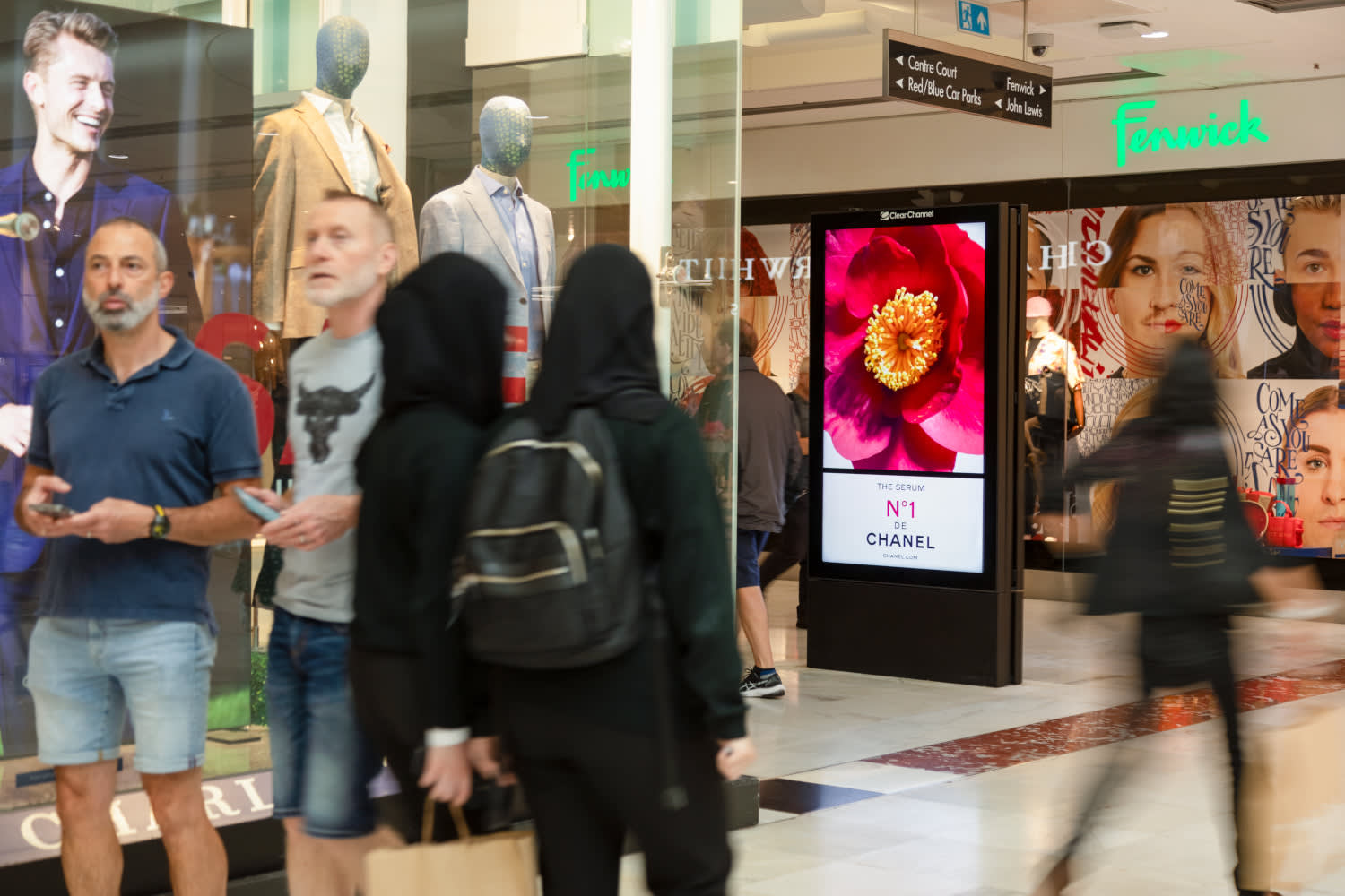 A digital screen inside a shopping mall featuring Chanel ad, with shoppers walking past