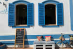 Two open windows with bright blue shutters on a white and blue striped building, with a chalkboard menu and colorful cushions.