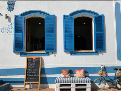 Two open windows with bright blue shutters on a white and blue striped building, with a chalkboard menu and colorful cushions.