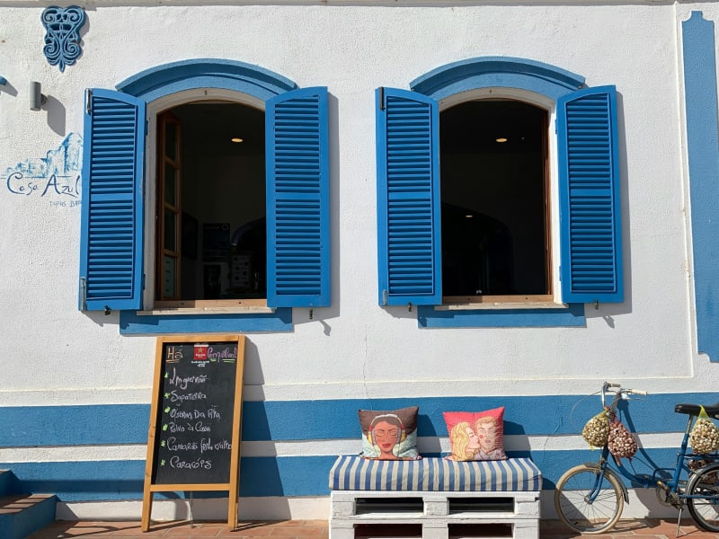 Two open windows with bright blue shutters on a white and blue striped building, with a chalkboard menu and colorful cushions.
