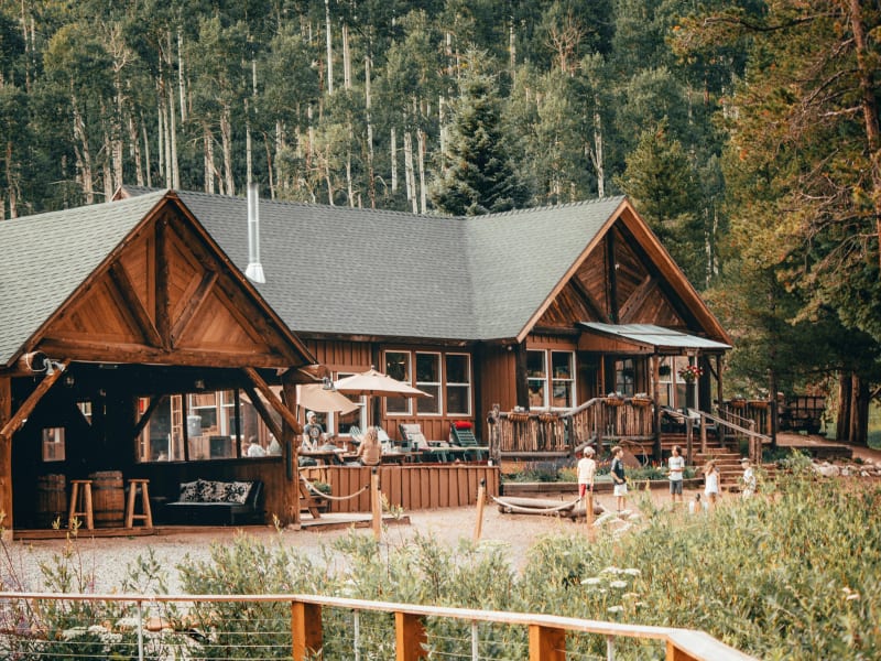 A large wooden lodge with a covered outdoor seating area and a deck, surrounded by trees.