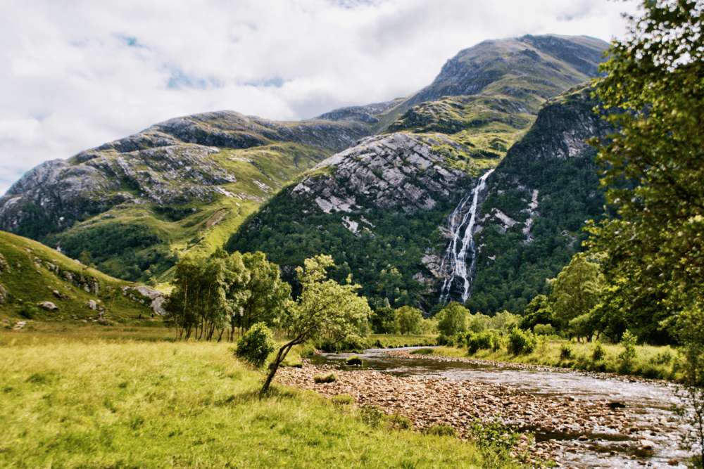 Grass covered mountains in the background, with grass and trees next to a river in the foreground.