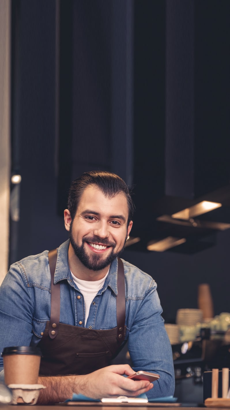 Smiling young craftsman wearing an apron sits and holds a mobile phone in his hands.
