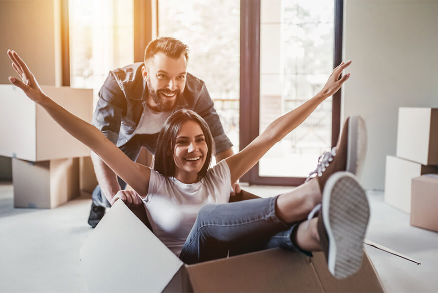 A young man pushes a girl in a large cardboard box around the room.