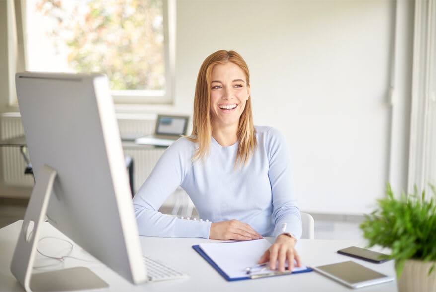 A smiling young woman sits at a desk and looks into the distance.