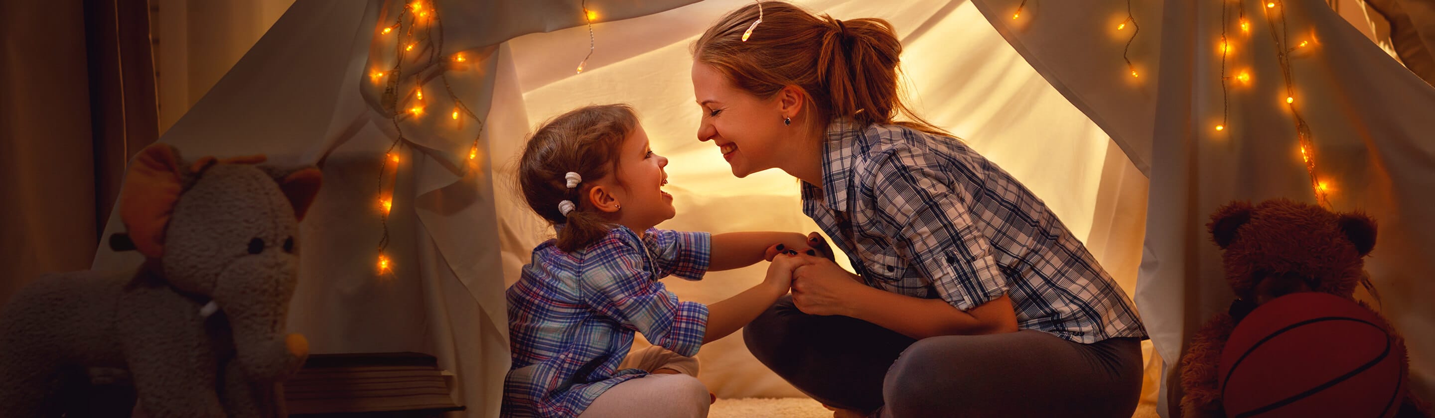 Smiling mother and girl sit in a tent in a children's room, holding hands.