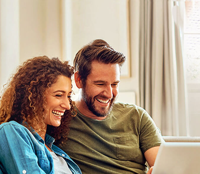 A smiling couple sits on a couch and looks at a laptop.