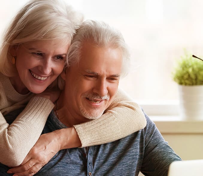 An elderly man looks at a laptop while an elderly woman hugs him from behind.
