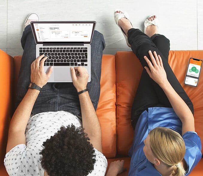 A young couple sits on an orange couch and looks at a laptop.
