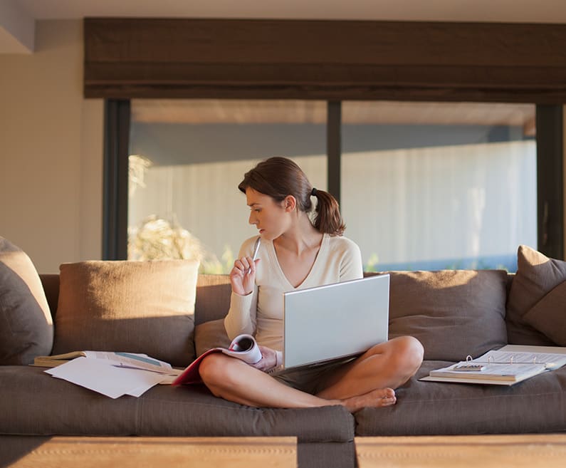 Woman on a couch with a laptop surrounded by papers.