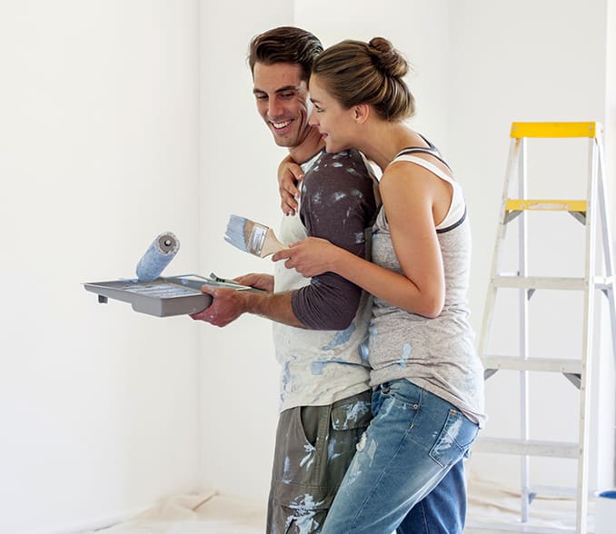 A young couple in an embrace holds wall painting kits in their hands.