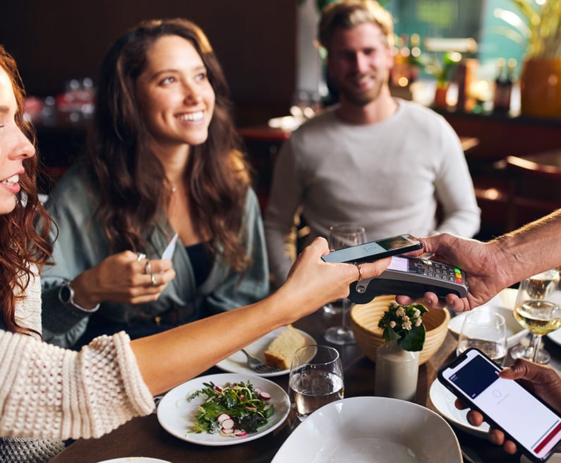 A young woman, accompanied by friends, holds her cell phone up to a POS device in a restaurant.