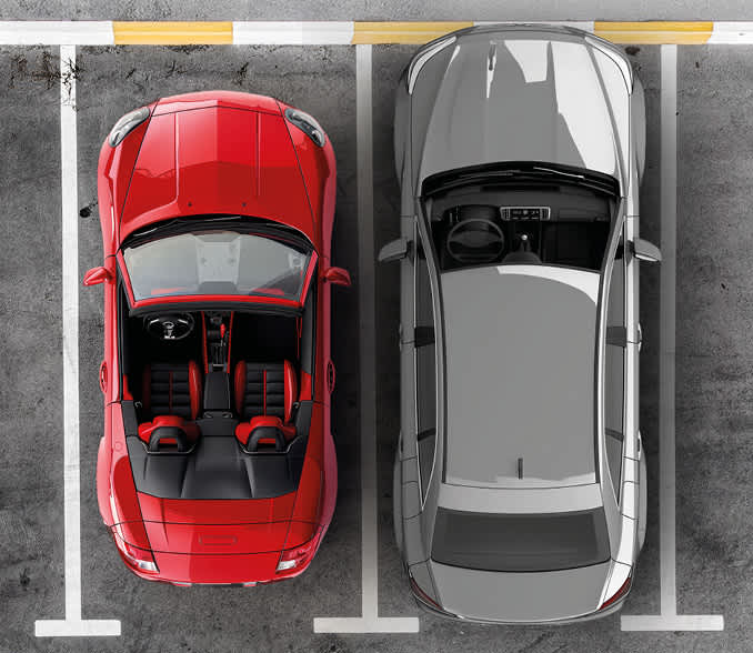 A red and gray car in a parking lot within the marked parking spaces.