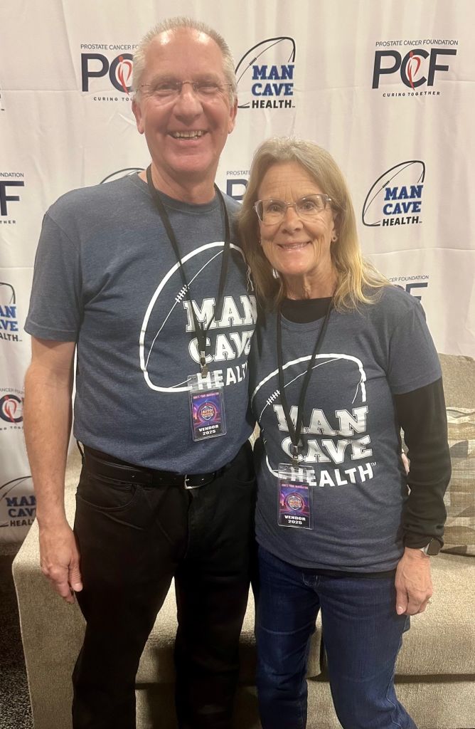 Ron stands next to his wife Nancy. They both are wearing glasses and they are wearing matching Man Cave Health shirts and are standing in front of a Man Cave Health x PCF banner.