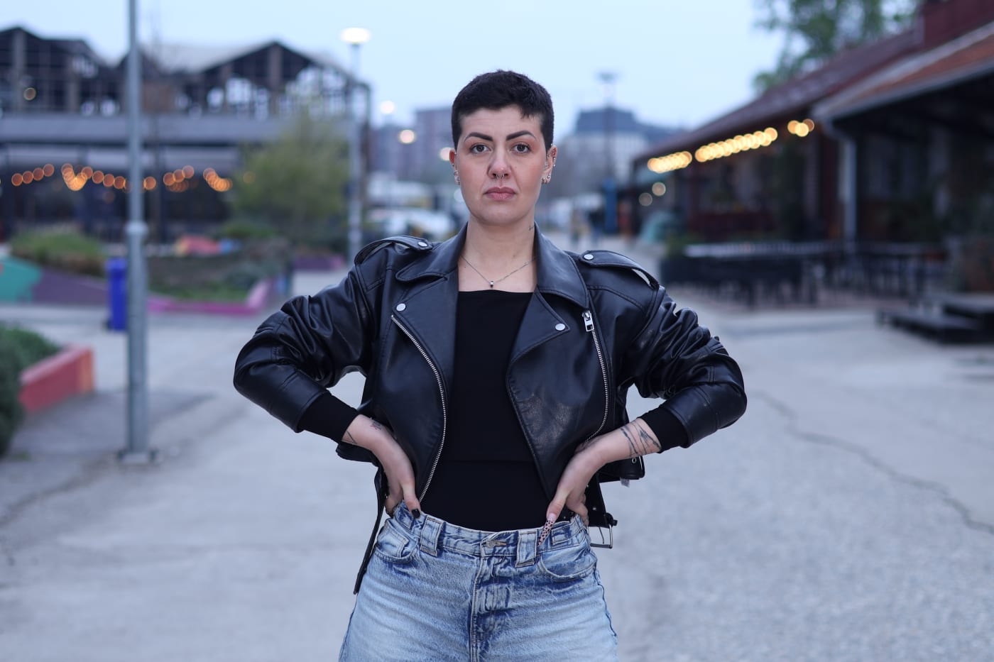 Portrait at dusk, hands on hips in a black leather jacket, city lights glowing behind